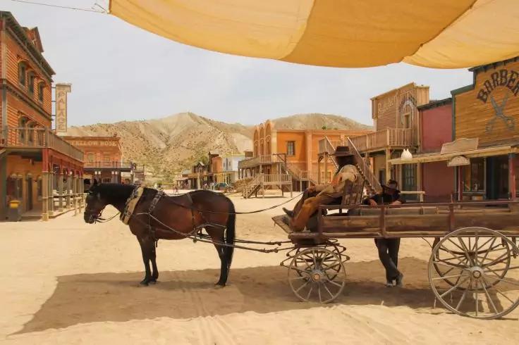 Zwei Pferde an einem h&ouml;lzernen Wagen auf einer sandigen Stra&szlig;e; h&ouml;lzerne Gesch&auml;ftsfronten links und rechts; niedrige Berge im Hintergrund; eine Person liegt auf dem Wagen; Sonnensegel im Vordergrund