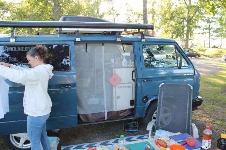 Blauer Camper mit ge&ouml;ffneter Schiebet&uuml;r, Person in wei&szlig;em Hoodie h&auml;lt ein Kleidungsst&uuml;ck, Campingtisch mit Tellern und Flaschen im Vordergrund, Koffer oder Paneel lehnt an der T&uuml;r, B&auml;ume im Hintergrund