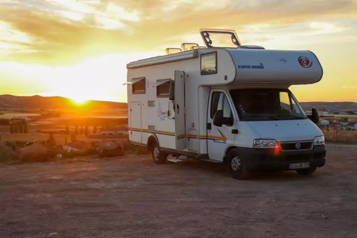 Wei&szlig;es Wohnmobil auf unbefestigtem Boden, Sonne tief am Horizont und leicht bew&ouml;lkter Himmel
