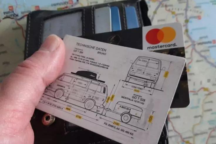 Hand h&auml;lt eine Karte mit technischen Zeichnungen eines Lieferwagens; im Hintergrund ein ge&ouml;ffnetes Portemonnaie mit einer Mastercard und eine Landkarte