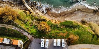 Luftaufnahme eines K&uuml;stenabschnitts mit Kiesstrand und Wellen, gr&uuml;ner Uferstreifen mit B&uuml;schen, mehrere wei&szlig;e Wohnmobile auf einem Parkplatz neben einem Geb&auml;ude