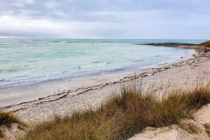 Sandstrand mit D&uuml;nengras im Vordergrund, t&uuml;rkis-gr&uuml;nes Meer mit kleinen Wellen, bew&ouml;lkter Himmel, rechts steinige K&uuml;stenbefestigung