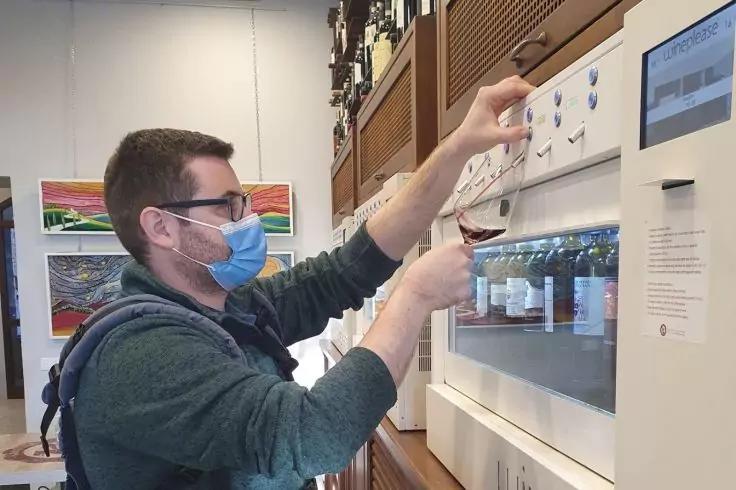 Person mit Brille und Mund-Nasen-Schutz entnimmt eine Flasche aus einem Wandger&auml;t mit mehreren Flaschen hinter Glas
