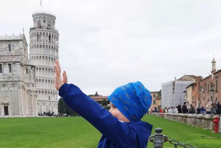 Person in blauem Mantel und blauer M&uuml;tze hebt den Arm vor dem Schiefen Turm von Pisa auf einer Rasenfl&auml;che; im Hintergrund Geb&auml;ude und Menschen