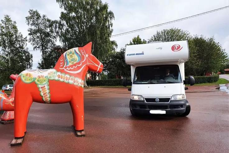 Gro&szlig;e rote Pferdeskulptur mit bunten Mustern links; wei&szlig;es Wohnmobil rechts auf nassem rotbraunem Asphalt; B&auml;ume und bew&ouml;lkter Himmel im Hintergrund