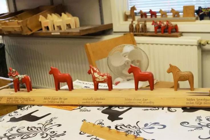 Mehrere kleine Holzpferde in Rot und Naturholz auf einem Tisch; im Vordergrund ein Holzlineal mit Schrift und eine wei&szlig;e Tischdecke mit schwarzem Blumenmuster; im Hintergrund ein Stuhl, ein kleiner Ventilator und weitere Holzpferde auf der Fensterban