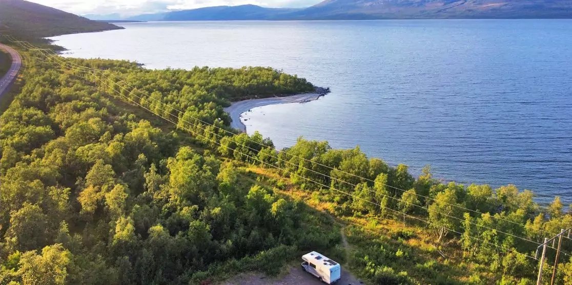 Luftaufnahme eines wei&szlig;en Wohnmobils am Waldrand neben einem See, Berge im Hintergrund, Stra&szlig;e links
