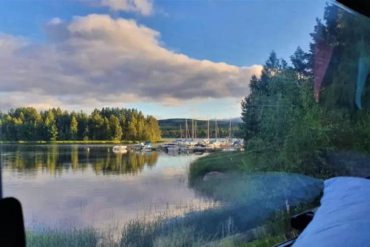 Blick durch ein Fenster auf einen See mit Schilf im Vordergrund, Boote und Segelyachten an einem kleinen Steg, Ufer mit B&auml;umen, Wolken am Himmel, Fensterrahmen und Kissen am rechten Bildrand