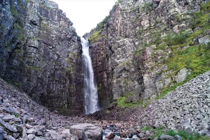 Wasserfall st&uuml;rzt zwischen hohen Felsw&auml;nden auf einen felsigen Boden; Ger&ouml;llhalde rechts, vereinzelte Vegetation an den Klippen, zwei Personen am Fu&szlig; des Wasserfalls