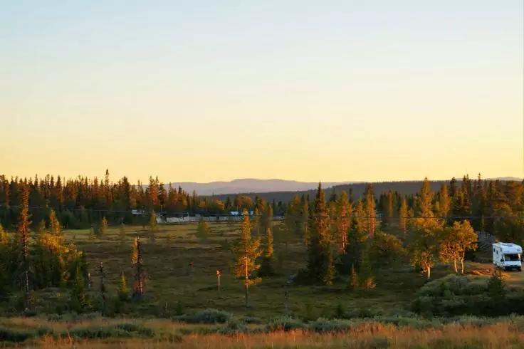 Offene grasbewachsene Ebene mit verstreuten Nadelb&auml;umen, kleiner See links, wei&szlig;es Wohnmobil rechts, entfernte H&uuml;gel am Horizont, gelb-orangefarbener Himmel