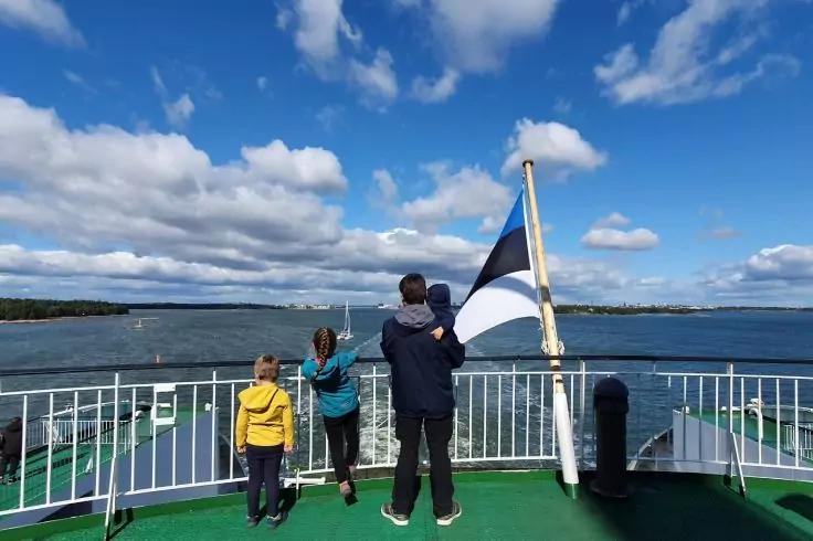 Drei Personen stehen mit dem R&uuml;cken zur Kamera auf dem Deck einer F&auml;hre; wei&szlig;e Reling, Flagge in Blau, Schwarz und Wei&szlig; an einem Mast, offenes Wasser und wolkiger Himmel