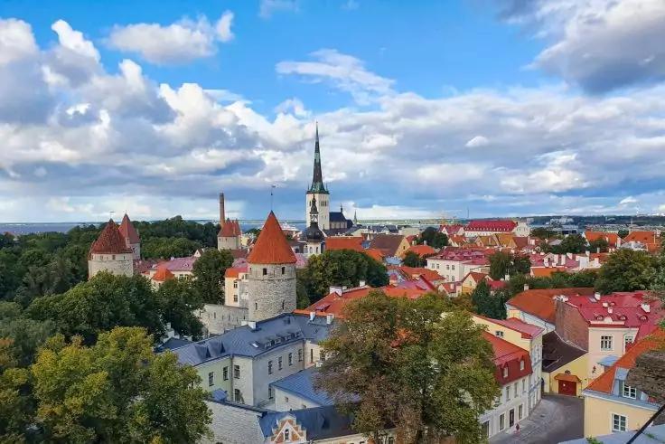 Blick auf eine Stadt mit roten D&auml;chern, einem hohen Kirchturm, runden steinernen T&uuml;rmen, B&auml;umen, wolkigem Himmel und Wasser am Horizont