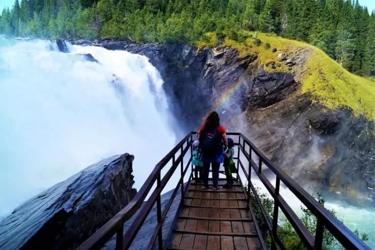 Zwei Personen von hinten mit Jacken und Rucks&auml;cken stehen auf einem h&ouml;lzernen Aussichtssteg vor einem gro&szlig;en Wasserfall mit Spr&uuml;hnebel, Felsen und Nadelwald im Hintergrund