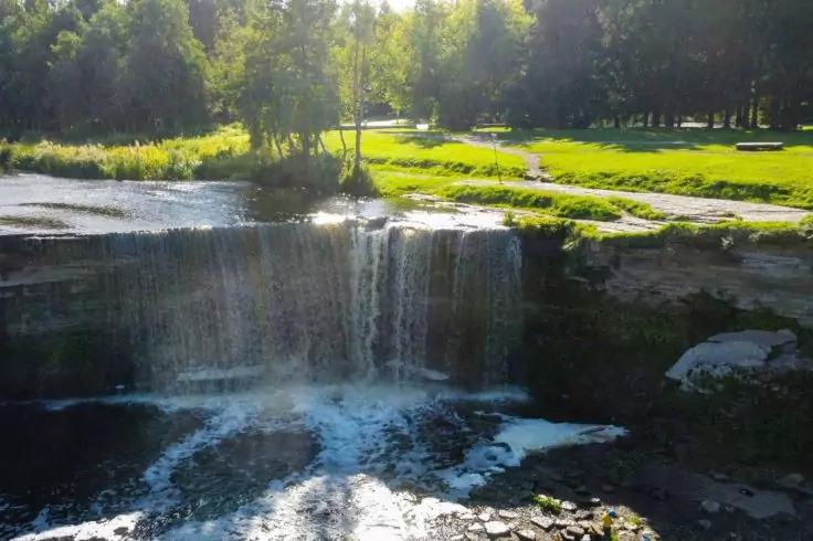 Kleiner Wasserfall &uuml;ber Felsen in einem Fluss; vorne sprudelndes Wasser, im Hintergrund gr&uuml;ner Rasen, B&auml;ume und ein Fu&szlig;weg