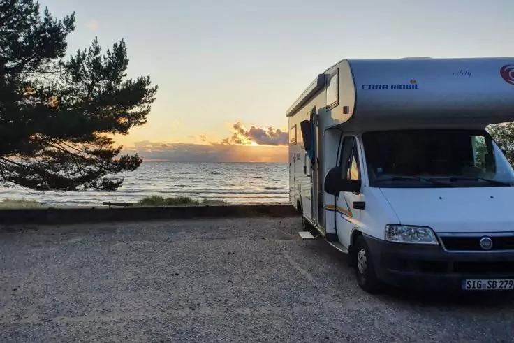 Wei&szlig;es Wohnmobil rechts auf einem Parkplatz am Meer, Wasser und Sonne am Horizont, Wolken im Himmel, Baum links