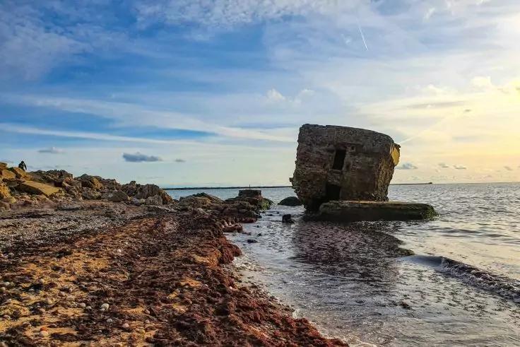 Schr&auml;g stehender gro&szlig;er Betonblock auf Felsen am Meer, felsiger Strand mit Algen, ruhiges Wasser und wolkiger Himmel bei niedrig stehender Sonne