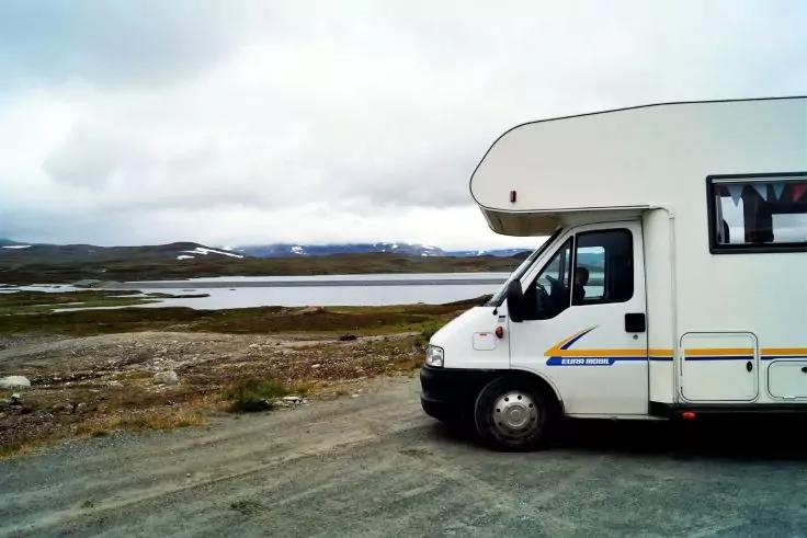 Vorderer rechter Teil eines wei&szlig;en Wohnmobils auf Schotter; im Hintergrund ein See, flaches Grasland und niedrige Berge unter wolkigem Himmel