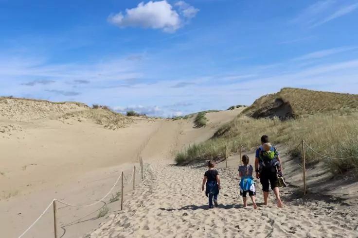 Drei Personen von hinten gehen auf einem sandigen Pfad zwischen D&uuml;nen; eine tr&auml;gt einen Rucksack; blauer Himmel mit einer Wolke