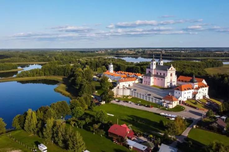 Luftaufnahme eines gro&szlig;en hellrosa Geb&auml;udes mit zwei T&uuml;rmen und roten D&auml;chern neben einem See, umgeben von B&auml;umen, Wiesen und blauem Himmel