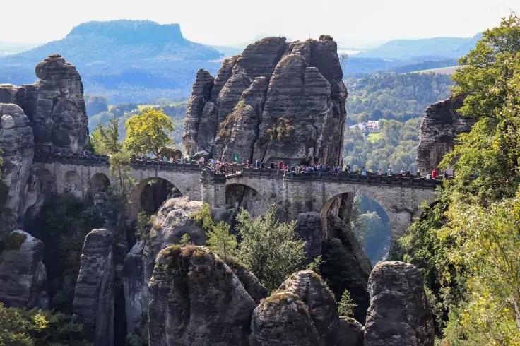 Steinerne Bogenbr&uuml;cke &uuml;ber hohe Felsnadeln, Menschen auf der Br&uuml;cke, bewaldete H&uuml;gel im Hintergrund