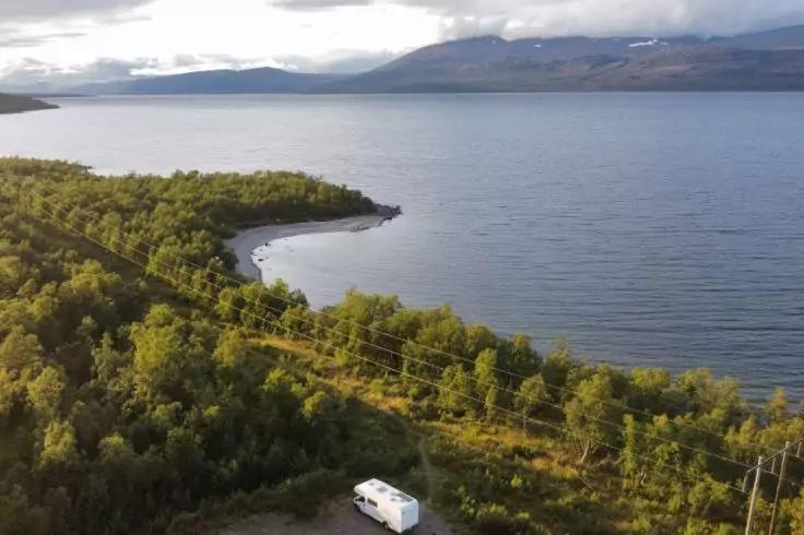 Ufer eines gro&szlig;en Sees mit bewaldeten H&auml;ngen, geschwungener Kiesstrand, wei&szlig;er Wohnwagen am Stra&szlig;enrand, Strommasten, Berge im Hintergrund und bew&ouml;lkter Himmel