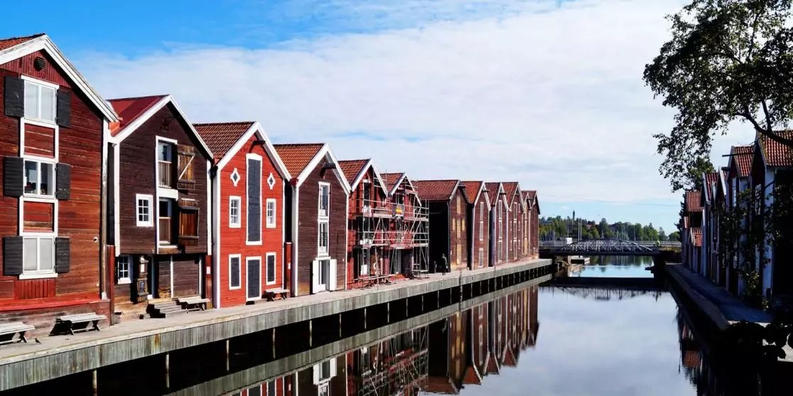 Rote h&ouml;lzerne H&auml;userreihe am Ufer eines Kanals, Spiegelung im Wasser, Br&uuml;cke im Hintergrund, blauer Himmel, Baum rechts