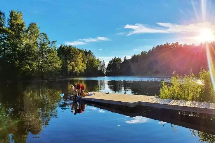 Zwei Personen sitzen am Ende eines Holzstegs an einem ruhigen See. Spiegelnde Wasseroberfl&auml;che, umgebende B&auml;ume, Schilf am Ufer, blauer Himmel mit Sonne und Sonnenstrahlen