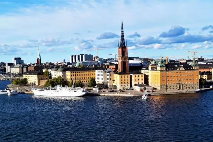 Blick auf ein Ufer mit Reihen von Geb&auml;uden, einem hohen Kirchturm mit Spitze, mehreren Schiffen im Wasser und blauem Himmel mit Wolken
