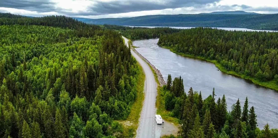 Luftaufnahme einer gewundenen Stra&szlig;e entlang eines Flusses, umgeben von dichtem Wald, wei&szlig;es Wohnmobil am Stra&szlig;enrand, bew&ouml;lkter Himmel