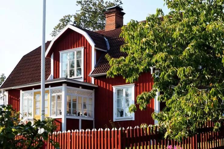 Rotes Holzhaus mit wei&szlig;en Fensterrahmen, verglaster Veranda, dunklem Dach, Schornstein, wei&szlig;em Lattenzaun und B&auml;umen im Vordergrund