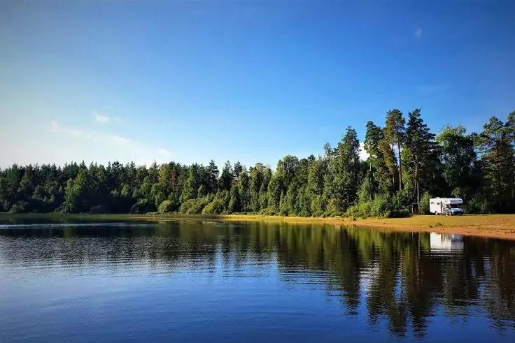 See mit spiegelnder Wasserfl&auml;che, sandigem Ufer, Baumreihe aus Nadel- und Laubb&auml;umen und rechts ein geparktes Wohnmobil unter blauem Himmel