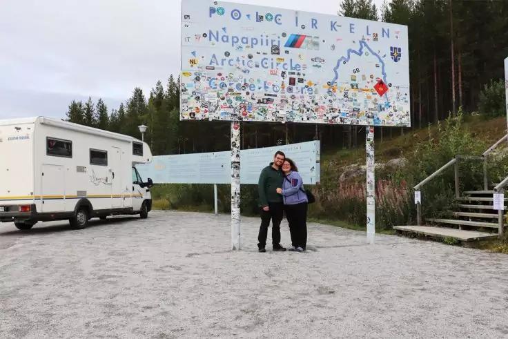 Zwei Personen stehen eng beieinander und umarmen sich vor einem gro&szlig;en Schild mit Karte und vielen Aufklebern; links ein Wohnmobil, rechts Holzstufen, im Hintergrund Nadelb&auml;ume