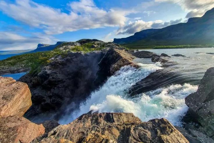 Wasserfall und Stromschnellen, die &uuml;ber steinige Felsen flie&szlig;en; Felsen im Vordergrund, Berge und bew&ouml;lkter Himmel im Hintergrund