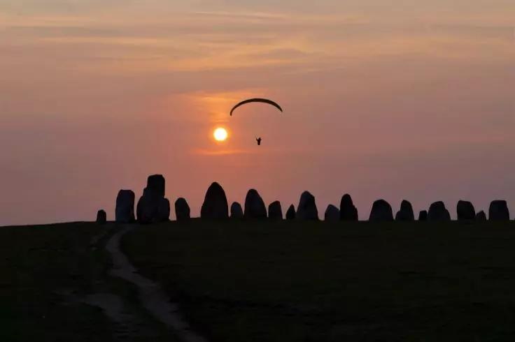 Silhouette eines Gleitschirmfliegers vor der Sonne knapp &uuml;ber dem Horizont, Reihe aufrecht stehender Steine und ein Pfad im Vordergrund