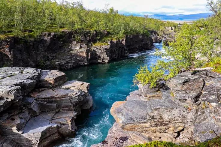 Felsklippen mit einem t&uuml;rkisfarbenen Fluss zwischen ihnen, Ufer mit gr&uuml;nen B&uuml;schen und B&auml;umen, blauer Himmel mit Wolken und entfernte H&uuml;gel