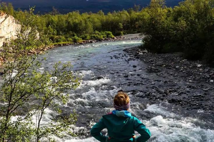 Person von hinten in t&uuml;rkisfarbener Jacke am Ufer eines Flusses mit Stromschnellen, umgeben von B&auml;umen, Felsen und Bergen unter bew&ouml;lktem Himmel