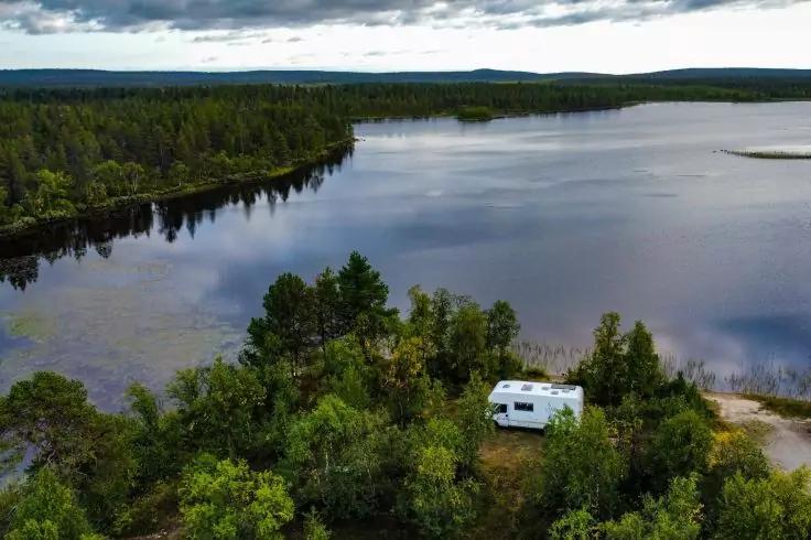 Wei&szlig;es Wohnmobil auf einer Lichtung am Ufer eines Sees, umgeben von gr&uuml;nem Wald; ruhige Wasserfl&auml;che mit Wolkenspiegelung