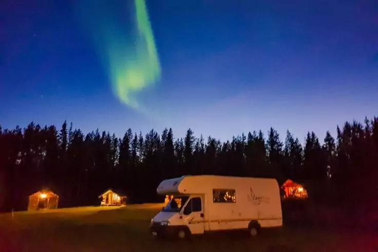 Wei&szlig;es Wohnmobil auf einem Grasfeld im Vordergrund rechts, drei beleuchtete Holzh&auml;user an der Waldrandlinie, dunkler Nadelwald, gr&uuml;nes Lichtband am dunkelblauen Nachthimmel