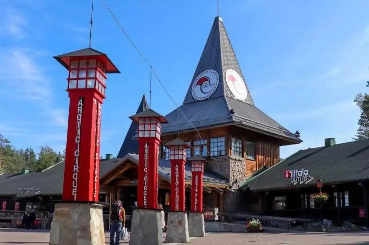 Gro&szlig;es Holzgeb&auml;ude mit spitzem Dach und rundem Emblem, davor mehrere rote S&auml;ulen mit wei&szlig;en Schildern, gepflasterter Platz mit Menschen, niedrige Gesch&auml;fte, blauer Himmel