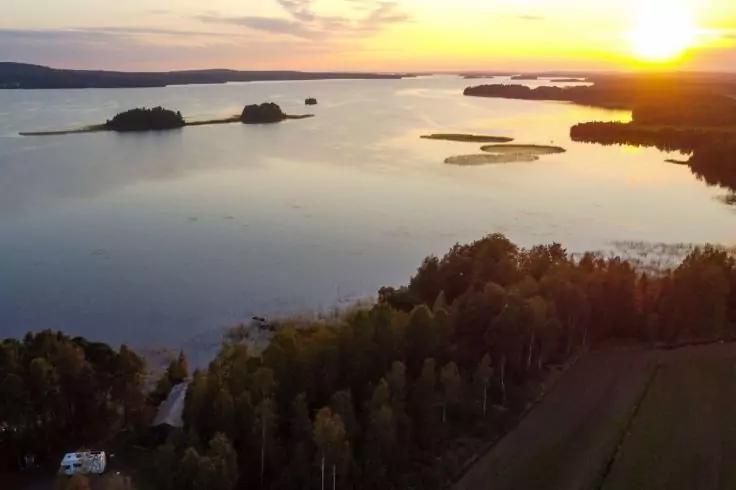 Luftaufnahme eines Sees mit kleinen Inseln, Ufer mit B&auml;umen, Feldern und einer Stra&szlig;e, ruhiges Wasser, Sonne nahe dem Horizont