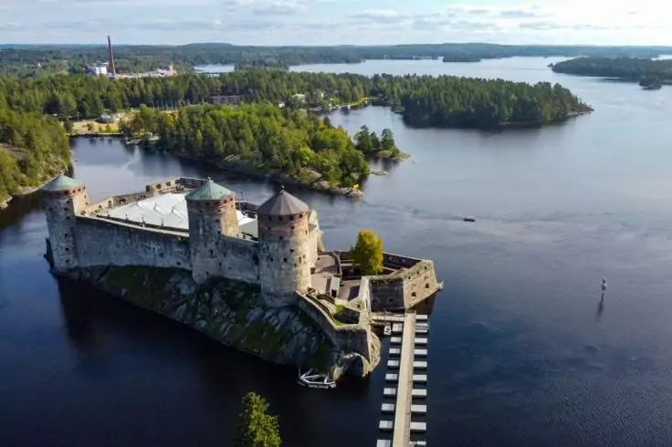 Steinbau mit drei runden T&uuml;rmen auf einer felsigen Insel, umgeben von einem See, mit Holzsteg und bewaldeten Uferinseln, wolkiger Himmel