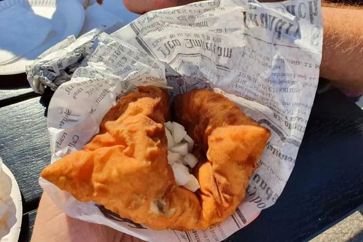 Zwei goldbraune, frittierte Teigtaschen mit wei&szlig;en Zwiebelst&uuml;cken, serviert auf Zeitungspapier in einer Papierschale; Hand und Teller am Rand sichtbar