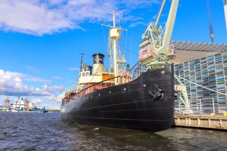 Gro&szlig;es schwarzes Schiff liegt am Kai neben einem Kran und einem Geb&auml;ude mit Glasfassade, Wasser im Vordergrund und blauer Himmel mit Wolken