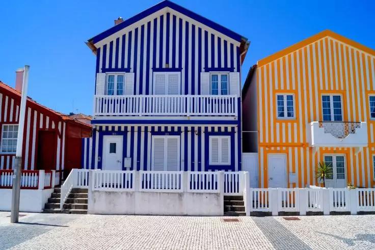 Drei zweist&ouml;ckige H&auml;user mit vertikalen Streifen (blau-wei&szlig; mittig, gelb-wei&szlig; rechts, rot-wei&szlig; links), wei&szlig;e Balkone und Treppen, gepflasterter Vorplatz und blauer Himmel