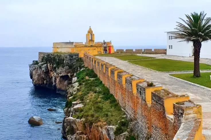 Klippenk&uuml;ste mit steinerner, gelb gestrichener Mauer und gepflastertem Weg, kleine Turmspitze mit Glocke am Ende, Meer links, Felsen im Wasser, Grasfl&auml;che, Palme und wei&szlig;es Geb&auml;ude rechts