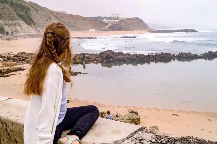 Person mit geflochtenem Haar sitzt auf einer niedrigen Mauer, R&uuml;cken zur Kamera, vor einem Sandstrand mit Felsen, flachen Gezeitenpools, Wellen und Klippen mit Geb&auml;uden unter bew&ouml;lktem Himmel