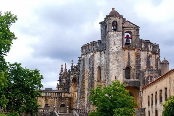 Gro&szlig;es steinernes Geb&auml;ude mit Glockenturm und sichtbarer Glocke, B&auml;ume im Vordergrund, bew&ouml;lkter Himmel
