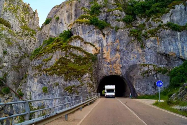 Wei&szlig;er Lkw an der Einfahrt eines Tunnels in einer felsigen, mit Vegetation bewachsenen Klippe; asphaltierte Stra&szlig;e mit Leitplanke und blauem, rundem Verkehrsschild rechts
