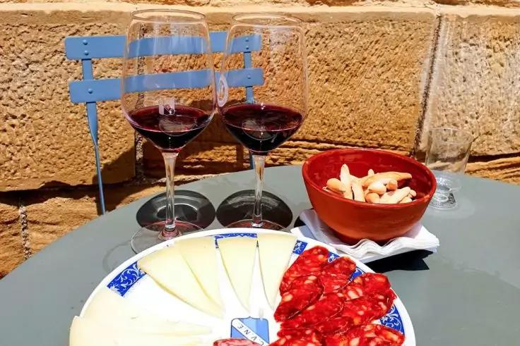 Zwei Gl&auml;ser Rotwein auf einem runden Tisch, Teller mit K&auml;sescheiben und roten Wurstscheiben, kleine Schale mit Brotst&uuml;cken, blauer Stuhl und Steinmauer im Hintergrund