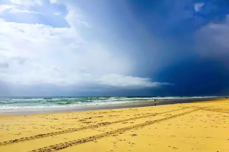 Sandstrand mit Reifenspuren, Wellen am Meer, eine Person am Ufer, heller Himmel links, dunkle Wolken rechts